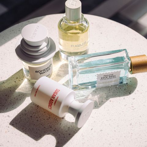 Quatre flacons de parfum différents sur une surface blanche mouchetée, éclairés par la lumière naturelle du soleil, chacun avec des formes et des étiquettes uniques.