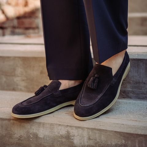 Personne portant des mocassins à glands en daim bleu marine avec des semelles crème, associés à un pantalon bleu marine, debout sur des marches en pierre.