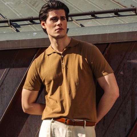 Un homme en polo marron et pantalon beige se tient à l'extérieur au soleil, une main dans la poche, devant une structure métallique.