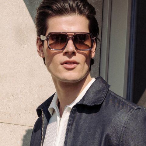 Un homme portant de grandes lunettes de soleil marron et une veste sombre se tient à l'extérieur au soleil et regarde légèrement sur le côté.