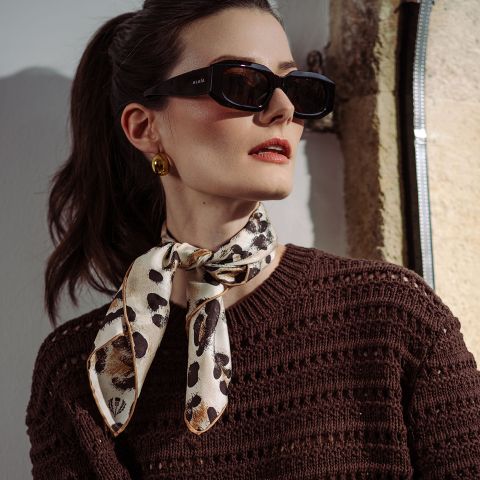 Une femme portant des lunettes de soleil noires, des boucles d'oreilles dorées, un haut en tricot marron et un foulard à motifs noué autour du cou, se tient au soleil.