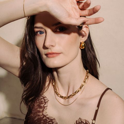 Une femme à la peau claire et aux cheveux bruns, portant des bijoux en or, pose avec un bras levé et regarde la caméra dans une douce lumière naturelle.