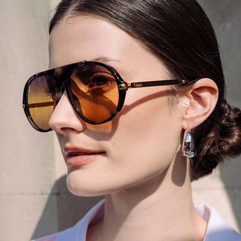 Femme aux cheveux foncés coiffés en un chignon bas, portant des lunettes de soleil marron surdimensionnées et des boucles d'oreilles argentées, se tenant devant un mur clair.