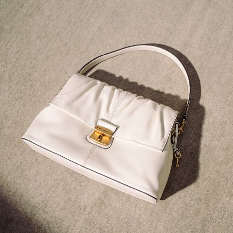 Sac à main crème avec boucle dorée et anse simple, posé sur une surface en béton clair à la lumière naturelle du soleil.