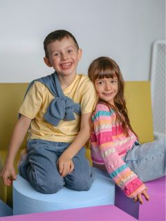 Deux enfants souriants sont assis sur des blocs de mousse colorés, le garçon en chemise jaune et jeans, la fille en pull rayé et jeans, et regardent la caméra.
