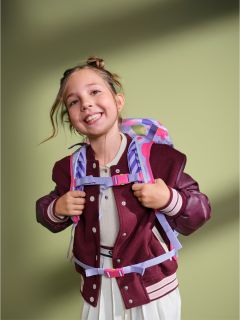 Une jeune fille souriante, vêtue d'une veste marron et d'une robe blanche, portant un sac à dos coloré, se tient devant un fond vert.