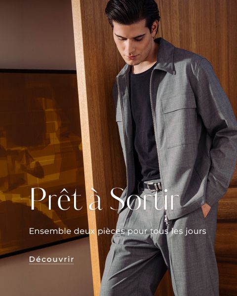 Un homme portant un élégant ensemble gris deux pièces sur une chemise foncée se tient à l'intérieur devant un panneau de bois. Un texte français fait la promotion de l'ensemble de vêtements.