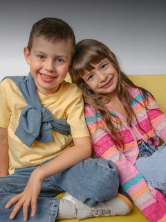 Un garçon souriant et une fille souriante sont assis l'un près de l'autre sur un canapé jaune. Le garçon porte une chemise jaune, la fille un pull à rayures multicolores.