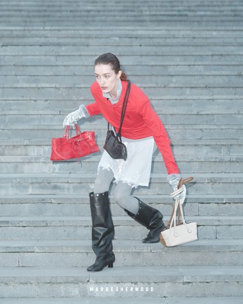 Une femme portant un haut rouge, des gants gris et des bottes noires descend un escalier en pierre, tenant un sac à main rouge et un sac à main beige, avec le texte "Margesherwood" en dessous.