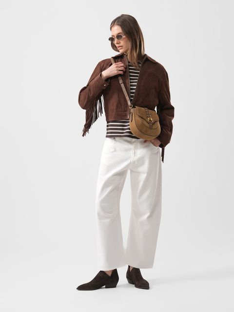 Femme en veste en daim marron à franges, chemise rayée, pantalon blanc à jambes larges, chaussures marron, lunettes de soleil et sac bandoulière marron clair, se tient là avec assurance.