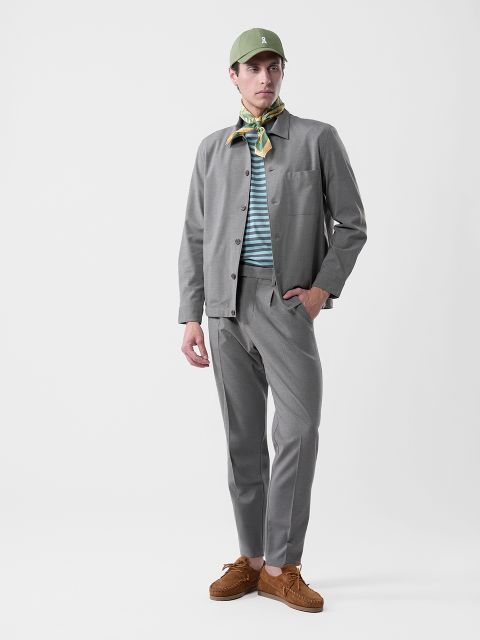 Homme en costume gris, chemise rayée, chapeau vert, foulard à motifs et chaussures marron devant un fond blanc uni.