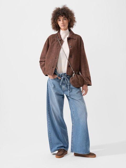 Personne portant une veste marron, un haut blanc, un jean bleu ample, des chaussures marron et un petit sac à main marron, se tenant devant un fond uni.