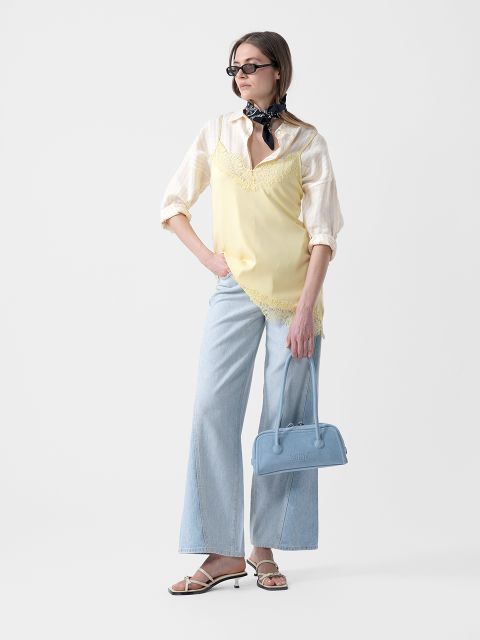 Femme portant des lunettes de soleil, un débardeur jaune sur une chemise rayée, un jean bleu clair à jambes larges, des sandales et un foulard, tenant un sac à main bleu.