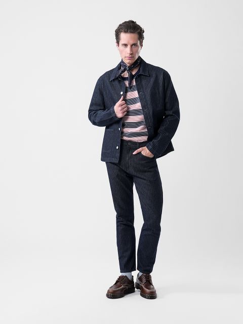 Un homme pose devant un fond blanc uni, vêtu d'une veste et d'un pantalon rayés bleu marine, d'une chemise rayée rose et grise, de chaussures marron et d'un foulard à motifs.