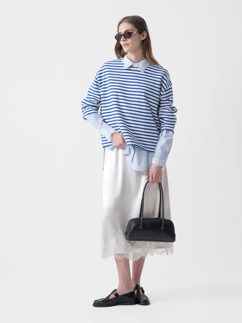 Femme portant des lunettes de soleil, un pull rayé bleu et blanc sur une chemise à col, une jupe blanche avec des finitions en dentelle, des chaussures basses noires et un sac à main noir.