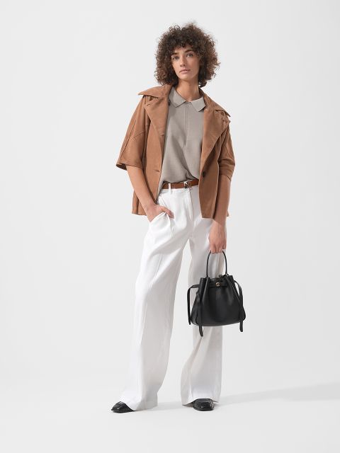 Une femme aux cheveux bouclés, portant une veste marron clair, une chemise beige et un pantalon blanc à jambes larges, tenant un sac à main noir, se tient devant un fond uni.