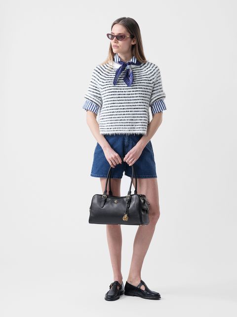 Femme portant des lunettes de soleil, un pull rayé sur une chemise à col, un short en jean et des chaussures noires, tenant un sac à main noir devant un fond blanc.