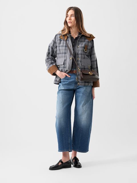 Une femme portant une veste à carreaux, un jean bleu à jambes larges, des chaussures noires et un sac à bandoulière marron se tient devant un fond blanc uni.