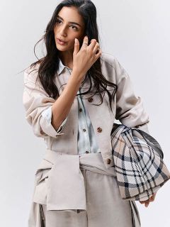 Une femme vêtue de vêtements beiges clairs avec un sac à carreaux, regardant sur le côté et touchant ses cheveux devant un fond uni.