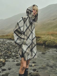 Une personne portant un imperméable à carreaux avec une capuche se tient près d'un ruisseau rocheux dans un paysage herbeux et brumeux, avec des collines en arrière-plan.