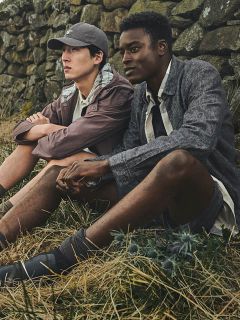 Deux hommes en tenue décontractée d'extérieur sont assis sur de l'herbe sèche, adossés à un mur de pierre, et regardent pensivement au loin.