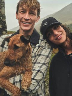 Un homme souriant tient un chien marron, tandis qu'il se tient à côté d'une femme souriante avec un bonnet noir, en extérieur par une journée nuageuse.