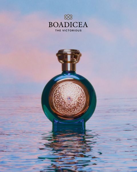 Un flacon de parfum bleu et or « Boadicea » repose sur une eau miroir devant un ciel pastel.
