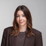 Une femme aux cheveux bruns ondulés, vêtue d'un blazer marron et de colliers superposés, pose devant un simple fond gris clair.