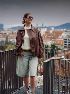 Une femme portant des lunettes de soleil et une veste marron se tient sur un balcon donnant sur un paysage urbain avec des toits rouges et des clochers par un jour nuageux.