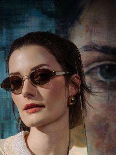 Femme aux cheveux bruns, portant des lunettes de soleil ovales et une boucle d'oreille dorée, se tient devant un portrait peint avec un grand œil et des nuances de bleu.