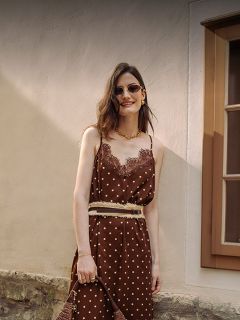 Une femme vêtue d'une robe marron à pois avec des garnitures en dentelle, des lunettes de soleil et des bijoux en or se tient souriante devant un mur beige et une fenêtre.