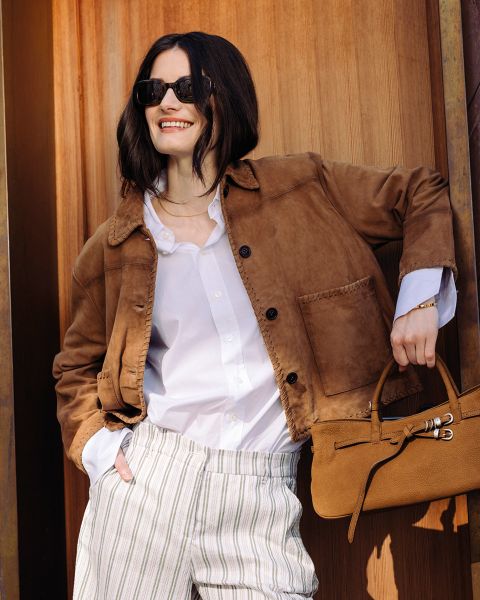 Femme avec des lunettes de soleil portant une veste marron, une chemise blanche et un pantalon rayé, souriant et tenant un sac à main marron devant un fond en bois.