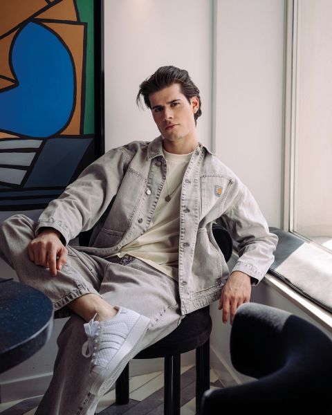 Un jeune homme en veste en jean claire et baskets blanches est assis confortablement sur une chaise près de la fenêtre dans une pièce moderne avec de l'art abstrait au mur.