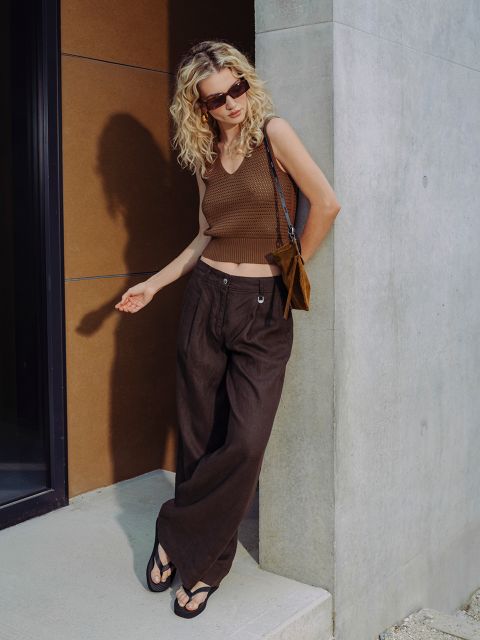 Une femme aux cheveux blonds bouclés porte des lunettes de soleil, un haut sans manches marron, un pantalon large marron et des sandales, et se tient devant un bâtiment moderne.