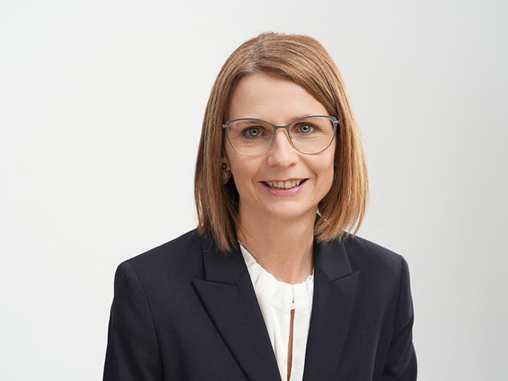 Femme aux cheveux châtain clair lisses, mi-longs, portant des lunettes, un blazer bleu marine et un chemisier blanc, souriant devant un fond clair et uni.