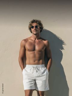 Un homme torse nu, portant des lunettes de soleil et un short rayé, se tient devant un mur ensoleillé, les mains dans les poches, et projette une ombre.