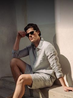 Un homme avec des lunettes de soleil, une chemise boutonnée gris clair et un short assorti est assis sur un escalier en béton au soleil devant un mur clair.