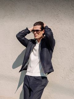 Un homme avec des lunettes de soleil, une veste foncée et un pantalon foncé se tient devant un mur texturé, les mains derrière la tête, et regarde au loin.