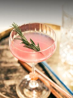 Un cocktail rose, garni d'une branche de romarin dans un verre à coupe, sur une surface en bois rustique avec des verres flous en arrière-plan.