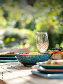 Un bol turquoise rempli de fruits est posé sur une table dressée avec des assiettes, du pain et un verre dans un jardin ensoleillé.