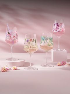 Quatre verres à vin avec des motifs floraux colorés, remplis de boissons, sont posés sur des podiums aux couleurs pastel avec de petites fleurs éparpillées sur un fond rose.
