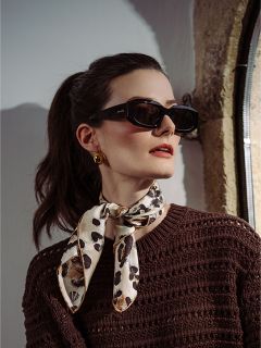 Femme aux cheveux foncés, coiffés en queue de cheval, portant des lunettes de soleil noires, des boucles d'oreilles dorées, un pull marron et un foulard à motif léopard, regardant sur le côté.