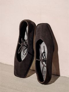 Une paire de chaussures en daim noir à bout carré avec des lacets, posée devant un mur clair et un sol clair.