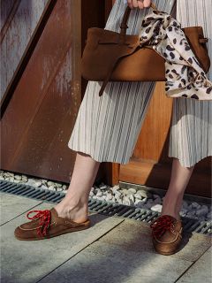 Personne portant un pantalon rayé à jambes larges et des mocassins marron avec des lacets rouges, tenant un sac à main marron avec un foulard à imprimé léopard noué à la poignée.