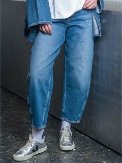 Une personne portant un jean bleu, une veste en jean, des chaussettes blanches et des baskets grises se tient dans une maison à côté d'une surface métallique.