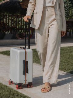Une personne vêtue d'un costume beige sort et tire une valise rigide blanche aux roues rouges et à la poignée noire sur un chemin pavé.