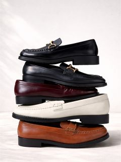 Une pile de quatre chaussons en cuir noir, bordeaux, blanc et marron, disposés verticalement devant un fond clair.