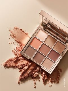 Une palette de fards à paupières avec neuf teintes dans des couleurs chaudes et neutres, avec un peu de poudre saupoudrée sur une surface beige.
