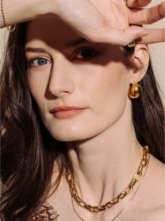 Une femme à la peau claire et aux cheveux bruns, portant des bijoux en or, pose avec sa main protégeant partiellement son visage de la lumière.