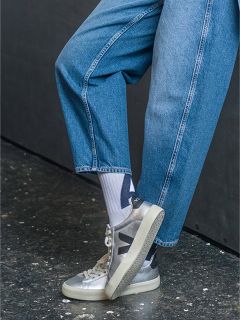 Personne en jean bleu, chaussettes blanches à motifs noirs et baskets Veja argentées, debout sur un sol foncé et tacheté, une jambe pliée.